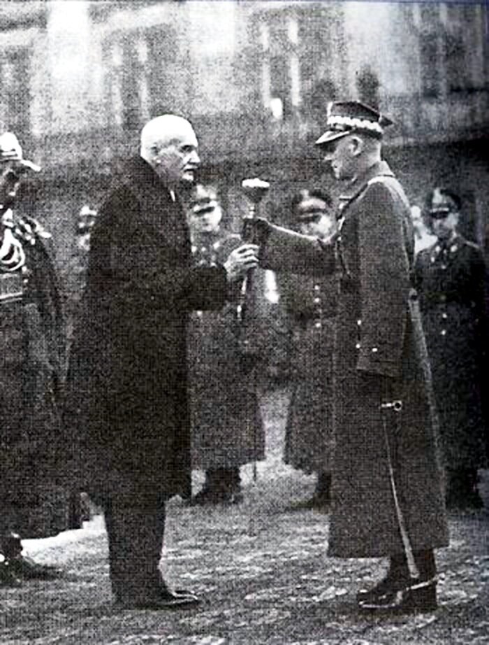 Na zdjęciu stoją dwaj mężczyźni. Po lewej stronie stoi prezydent Mościcki. Ubrany jest w długi płaszcz. Wręcza Edwardowi Rydz-Śmigłemu buławę marszałkowską. Edward Rydz-Śmigły ubrany jest w długi wojskowy płaszcz. Na głowie ma czapkę wojskową. W lewej ręce trzyma opuszczoną szablę. Prawą odbiera buławę marszałkowską. W tle widać pięciu żołnierzy. 