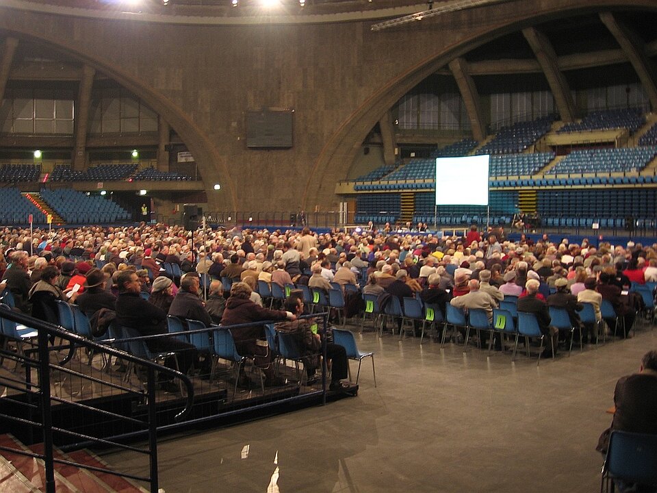 Zdjęcie przedstawia zgromadzenie ludzi w hali sportowej. Ludzie siedzą na krzesłach na płycie hali. W tle widoczny jest stół prezydialny, za którym siedzi kilka osób. 