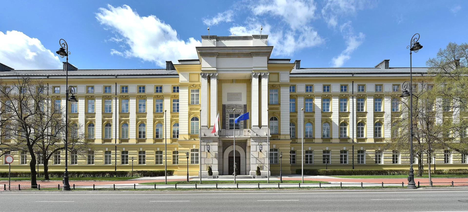 Fasada Kancelarii Prezesa Rady Ministrów w Warszawie. Monumentalny, jasny budynek w stylu klasycyzującym z kolumnowym portykiem - widoczny od strony Alej Ujazdowskich. Budynek otoczony zielenią i z elementami ogrodzenia w kadrze.
