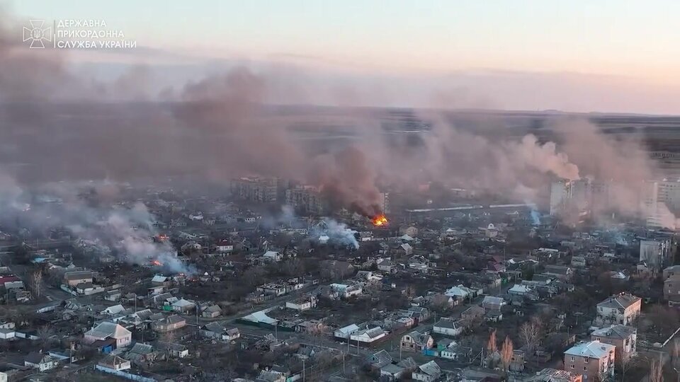 Zdjęcie przedstawia panoramę miasta Bakhmut podczas bitwy w 2023 roku; nad zabudowaniami widoczne są liczne kłęby dymu i ogniska pożarów, świadczące o zniszczeniach spowodowanych działaniami wojennymi. Na pierwszym planie znajdują się uszkodzone budynki mieszkalne i domy jednorodzinne, w tle widać większe bloki mieszkalne, a cała scena utrzymana jest w przygaszonych barwach. Zdjęcie ilustruje skalę zniszczeń infrastruktury oraz skutki konfliktu zbrojnego dla terenów miejskich.
