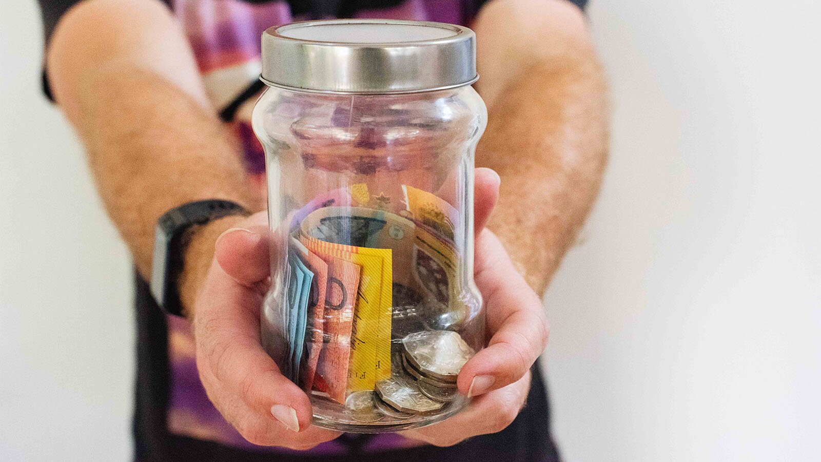 Zdjęcie przedstawia trzymany w męskich dłoniach szklany słoik z metalową nakrętką. W słoiku widoczne są banknoty o różnych nominałach oraz monety.
