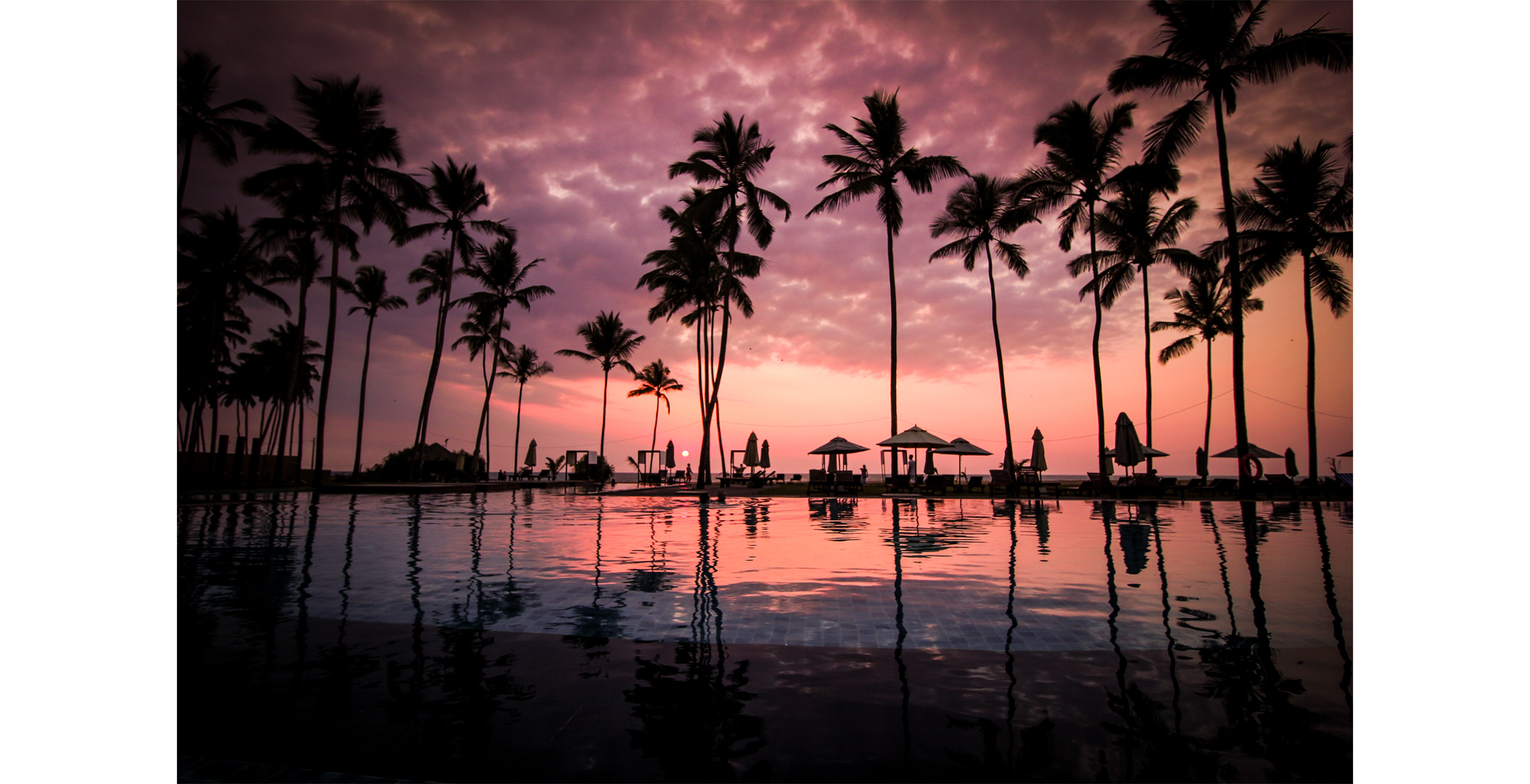 Na zdjęciu pokazany jest zachód słońca nad basenem. W wodzie odbijają się ciemne chmury i palmy rosnące wokół. W oddali widoczne są również parasole i leżaki.