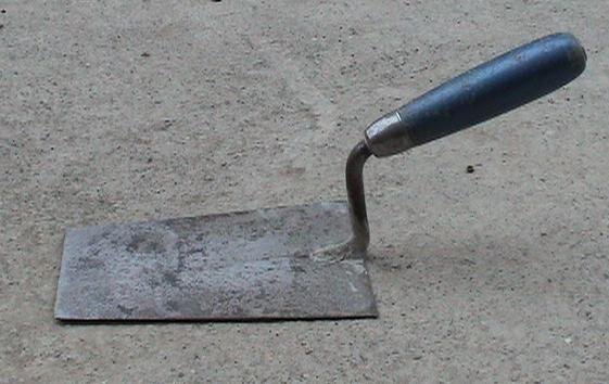 Zdjęcie przedstawia kielnię. Jest to płaskie narzędzie w kształcie trapezu z rączką.