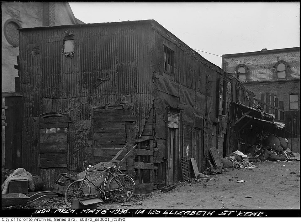 Zdjęcie przedstawia slumsy w Toronto. Na pierwszym planie znajduje się rower oparty o wózek do przewożenia rzeczy. Dalej stoi długi budynek zbudowany z blach oraz z zabitymi deskami otworami. Przed nim znajdują się śmieci.