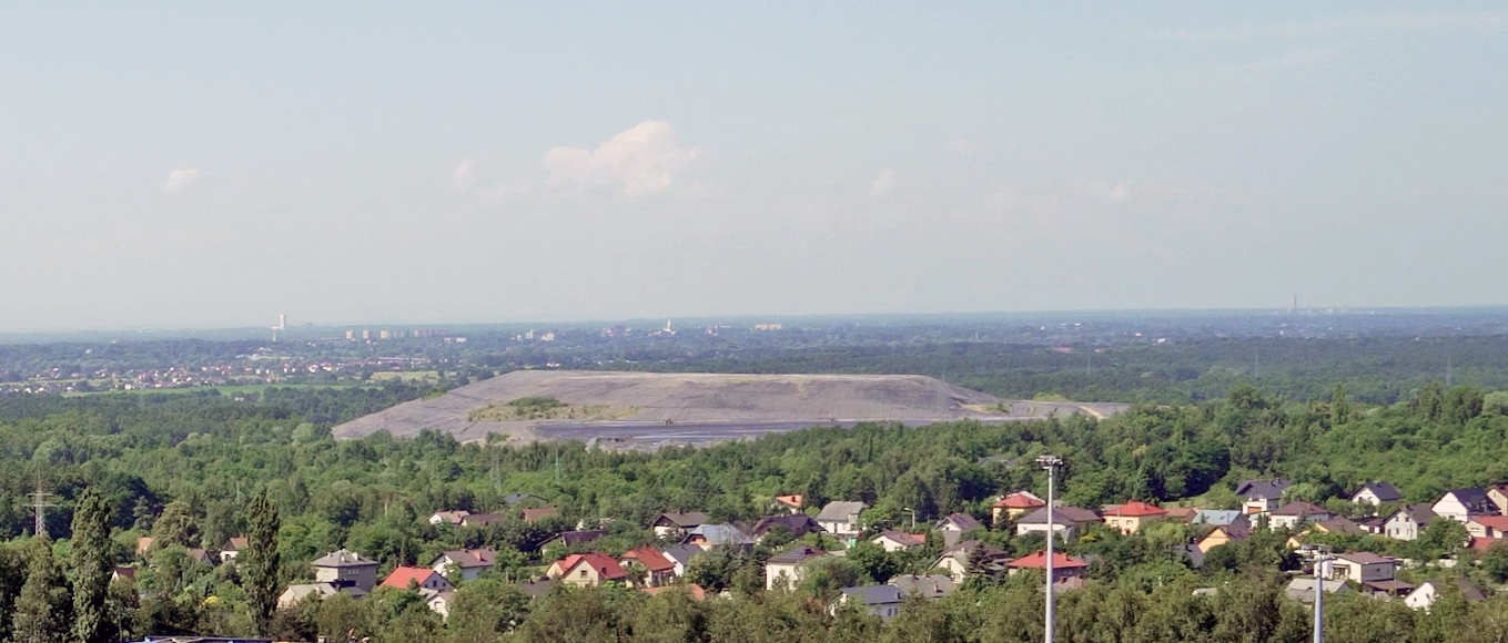 Na zdjęciu pokazano widok hałdy z lotu ptaka. Widoczna w centralnym punkcie zdjęcia hałdy jest o kształcie pryzmy. Pryzma góruje nad otoczeniem. W tle widać niskie zabudowania. Poniżej zdjęcia widoczny jest przycisk multimedialny z nagraniem audio. Treść nagrania jest zgodna z tytułem zdjęcia.