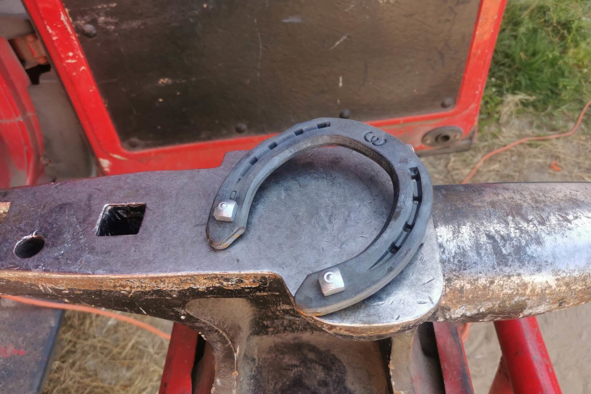 Zdjęcie przedstawia podkowę z hacelami. Jest to metalowy element wygięty w kształt litery U. Podkowa zwęża się na końcach. Po obu stronach podkowy widoczne są wgłębienia, w których znajdują się po cztery prostokątne otwory na gwoździe. Na górze podkowy widoczna jest sygnatura, a na końcach ramion hacele, czyli metalowe, wypukłe elementy w kształcie sześcianu.