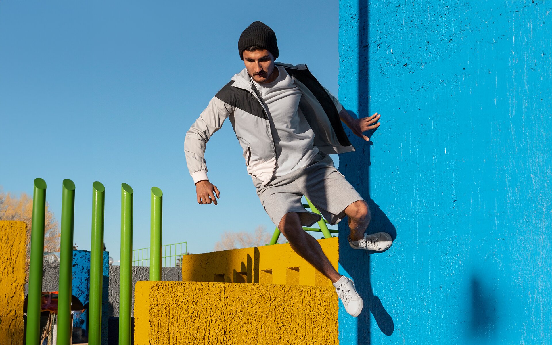Zdjęcie przedstawia mężczyznę wykonującego skok przy pionowej ścianie. Mężczyzna ubrany jest w koszulkę, na którą ma zarzuconą rozpiętą szarą bluzę, szare szorty, sportowe buty oraz czarną czapkę. Pokazany jest w locie. Lewą ręką i lewą nogą dotyka niebieskiej ściany. Zaś jego prawa ręka i noga wiszą w powietrzu. W tle widoczne są żółte murki i pięć zielonych słupków.