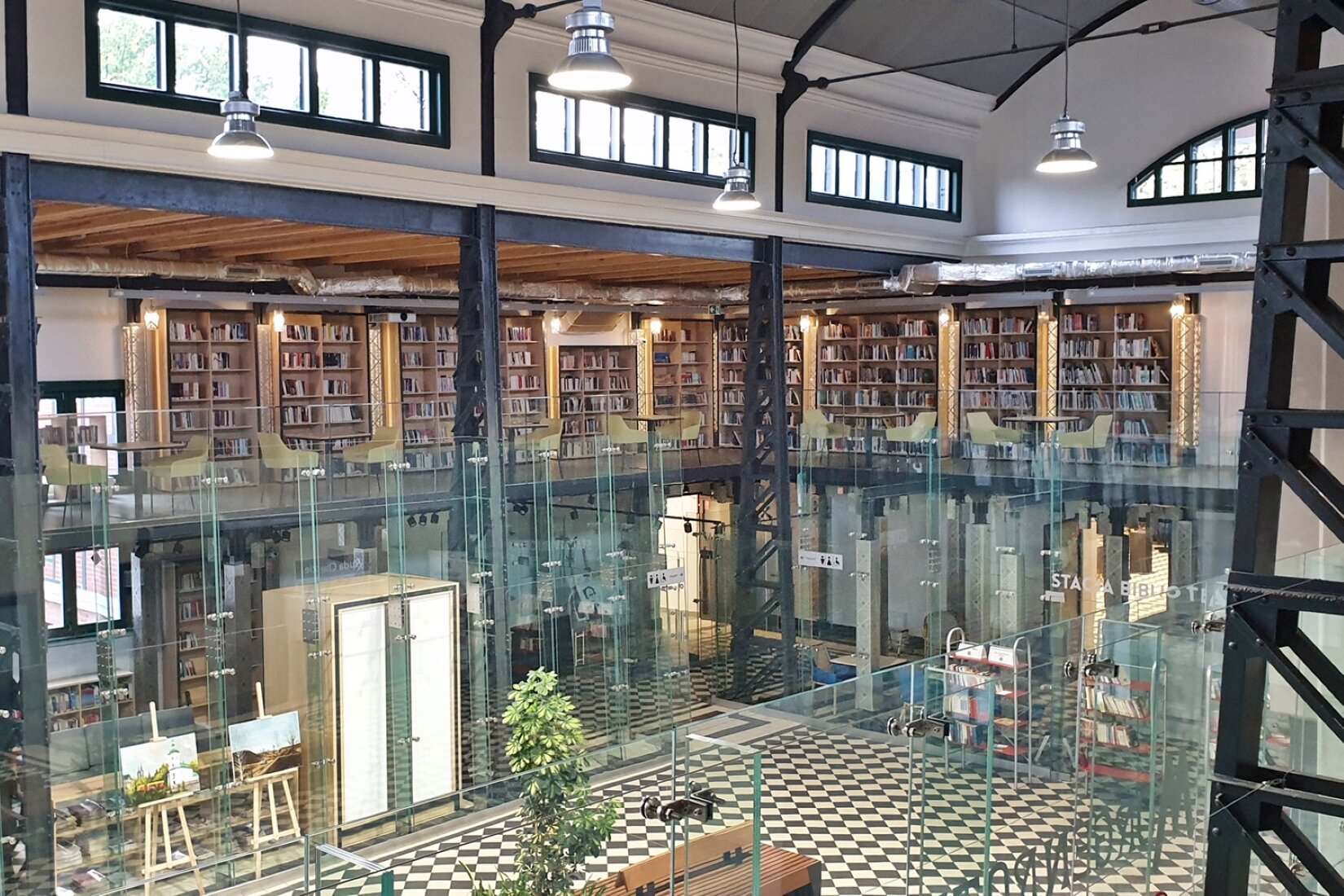 Na zdjęciu przedstawiono czytelnię z dwoma piętrami. Niższe piętro stanowi hol w którym stoją regały, szafa oraz dwie sztalugi, wyżej jest pomieszczenie w którym znajdują się regały z książkami. Obok nich stoją stoliki z żółtymi krzesłami.  Sufit jest podtrzymywany czarnymi, metalowymi stelażami. Wyżej znajdują się czarne, małe okna. Z sufitu zwisają srebrne lampy. 