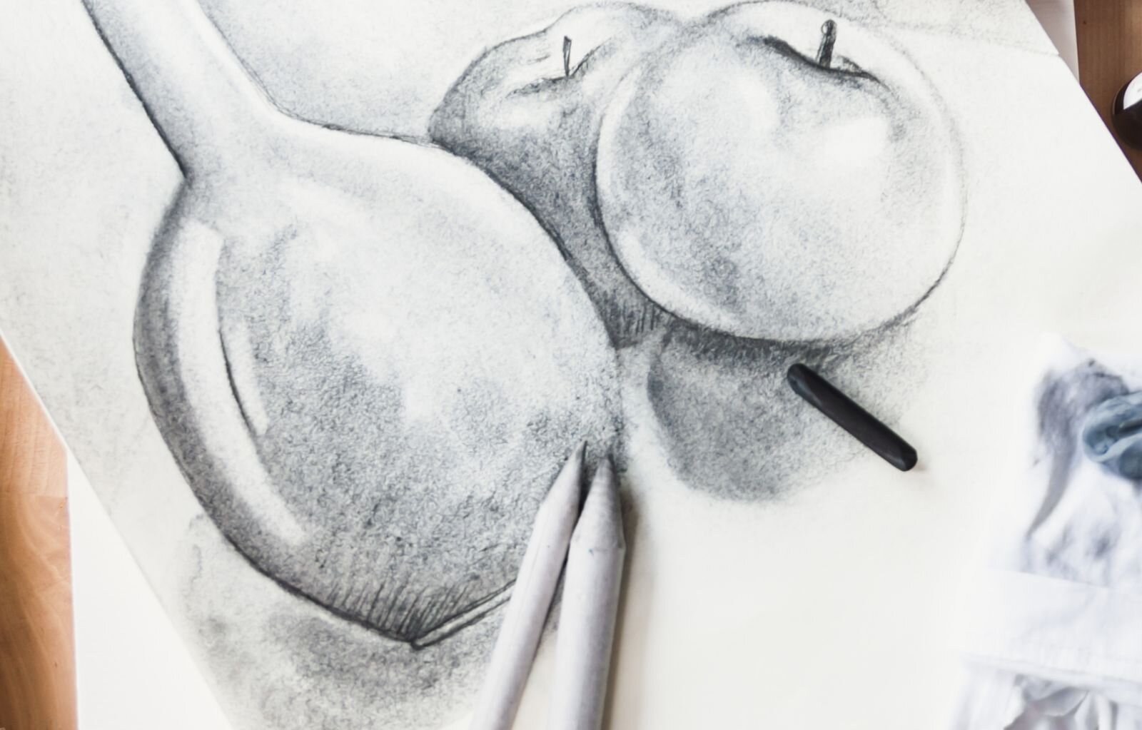 Rysunek węglem przedstawia wazon i 2 jabłka. Na rysunku obok jabłek leży węgiel, którym był wykonany rysunek, a na wazonie dwa ołówki, jeden grubszy od drugiego. Węgiel pozostawia miękki kontur, który roztarty tworzy płaszczyznę stołu i bryły przedmiotów. Na stole pojawia się cień jabłek i wazonu, a pociągnięcia gumki są jak muśnięcia światła.