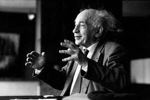 Na czarno‑białym zdjęciu znajduje się Alfred Sauvy. Jest to starszy mężczyzna o siwych rozczochranych włosach. Jest on ubrany w garnitur. Siedzi przy biurku i przemawia gestykulując. 