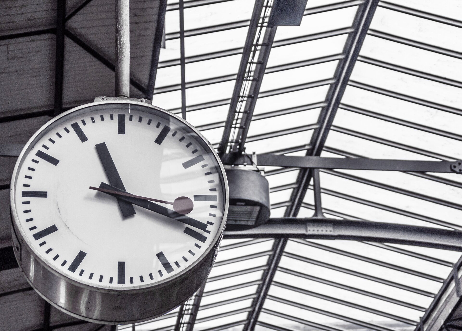 Na zdjęciu zegar wskazówkowy z sekundnikiem zakończonym kółkiem, umocowany na metalowych belkach pod szklanym sufitem hali. Zegar jest wykonany z blachy, na białej tarczy wskazówki pokazują godzinę 11.19 (i osiemnaście sekund). Na tarczy poszczególne godziny oznaczone są kreskami, a nie liczbami.