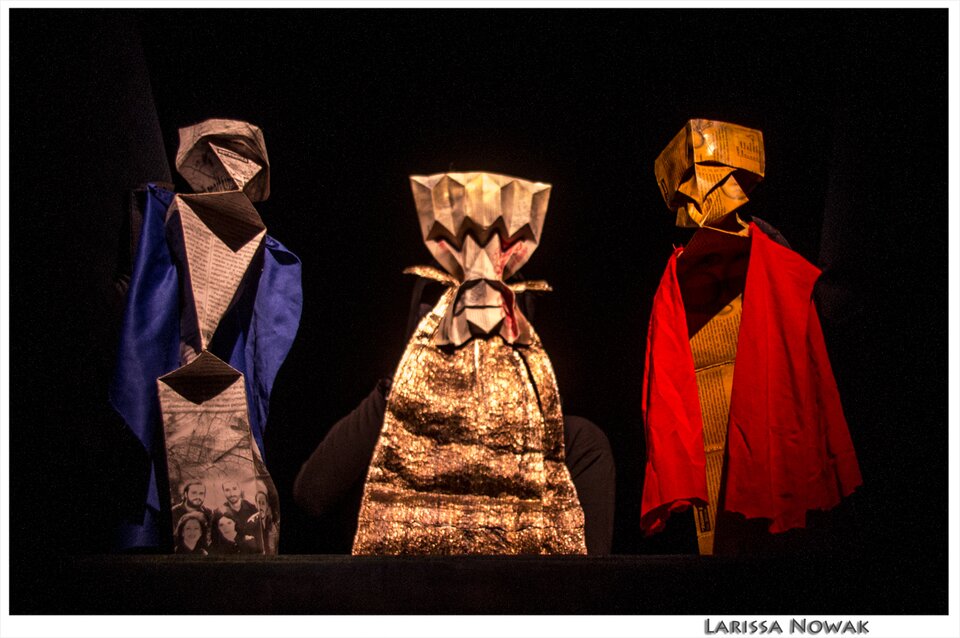 Ilustracja przedstawia dłonie w czarnych rękawiczkach trzymające trzy postacie stworzone z papieru. Postacie są schematyczne, to paski papieru rozszerzone ku dołowi, z zebranym na górze końcem, na którym doczepione są papierowe elementy imitujące głowy. Postać po lewej stronie ubrana jest w granatowy kawałek materiału, postac po prawo w czerwony kawałek materiału, który ma symbolizować płaszcz, środkowa postać jest złota.