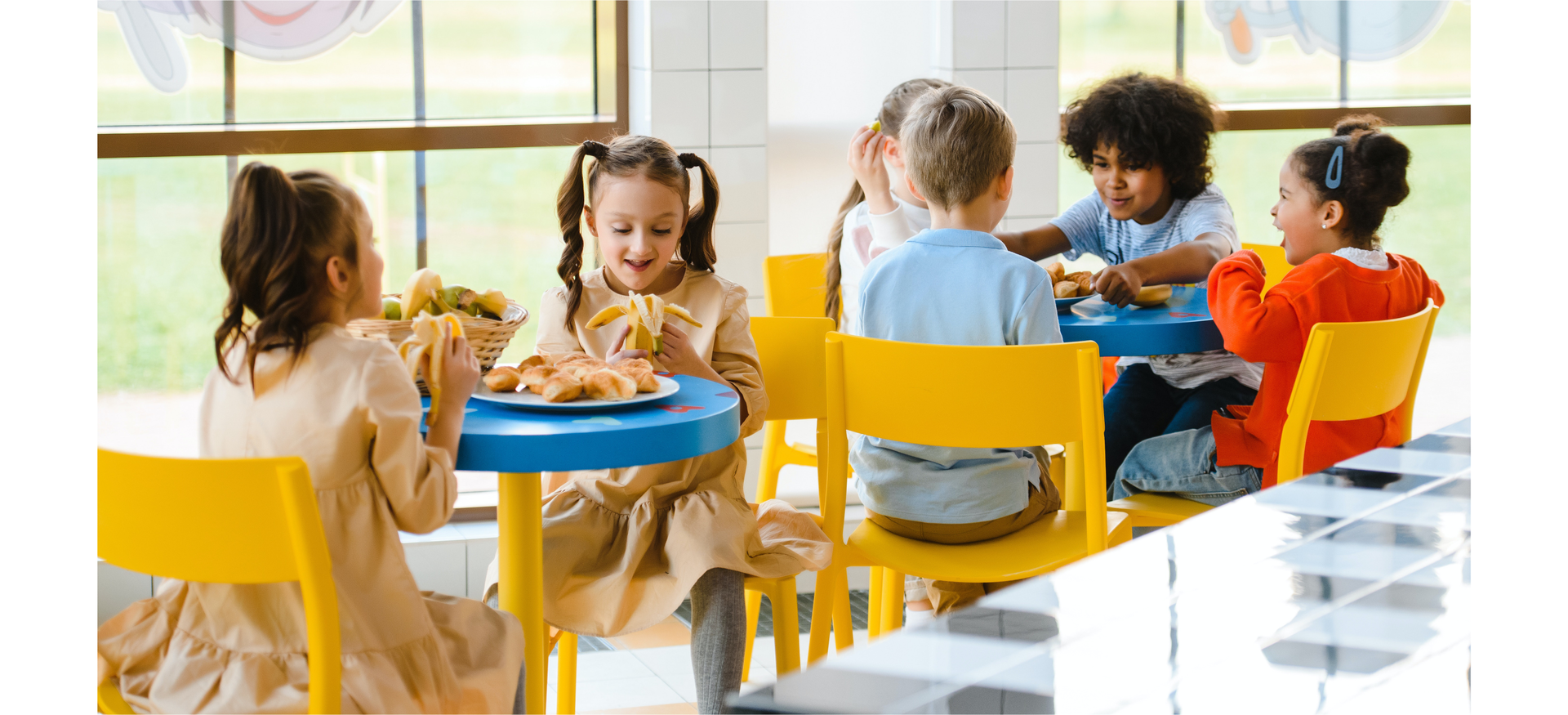 Zdjęcie przedstawia dzieci siedzące przy stolikach na stołówce. Na bliższym planie przy stoliku siedzą dwie dziewczynki, które jedzą banany. Na stole widoczne są owoce i ciasteczka. Na dalszym planie, przy drugim stoliku siedzi czworo dzieci, dwie dziewczynki i dwóch chłopców.