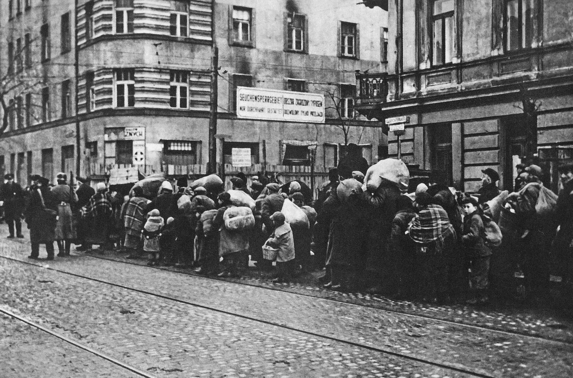 Czarno‑biała fotografia przedstawia ulicę centrum miasta, na której w rzędzie stoją ludzie trzymający liczne tobołki i pakunki. Na ulicy znajdują się tory tramwajowe. 