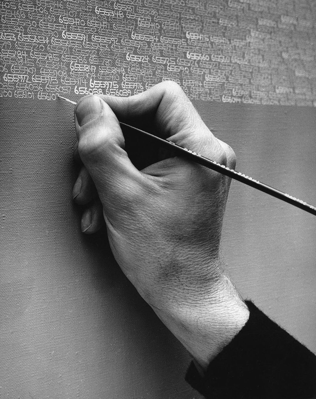 Fotografia dłoń artysty w czasie pracy nad swoim dziełem. Artysta za pomocą pędzla nanosi białe liczby na szare tło.