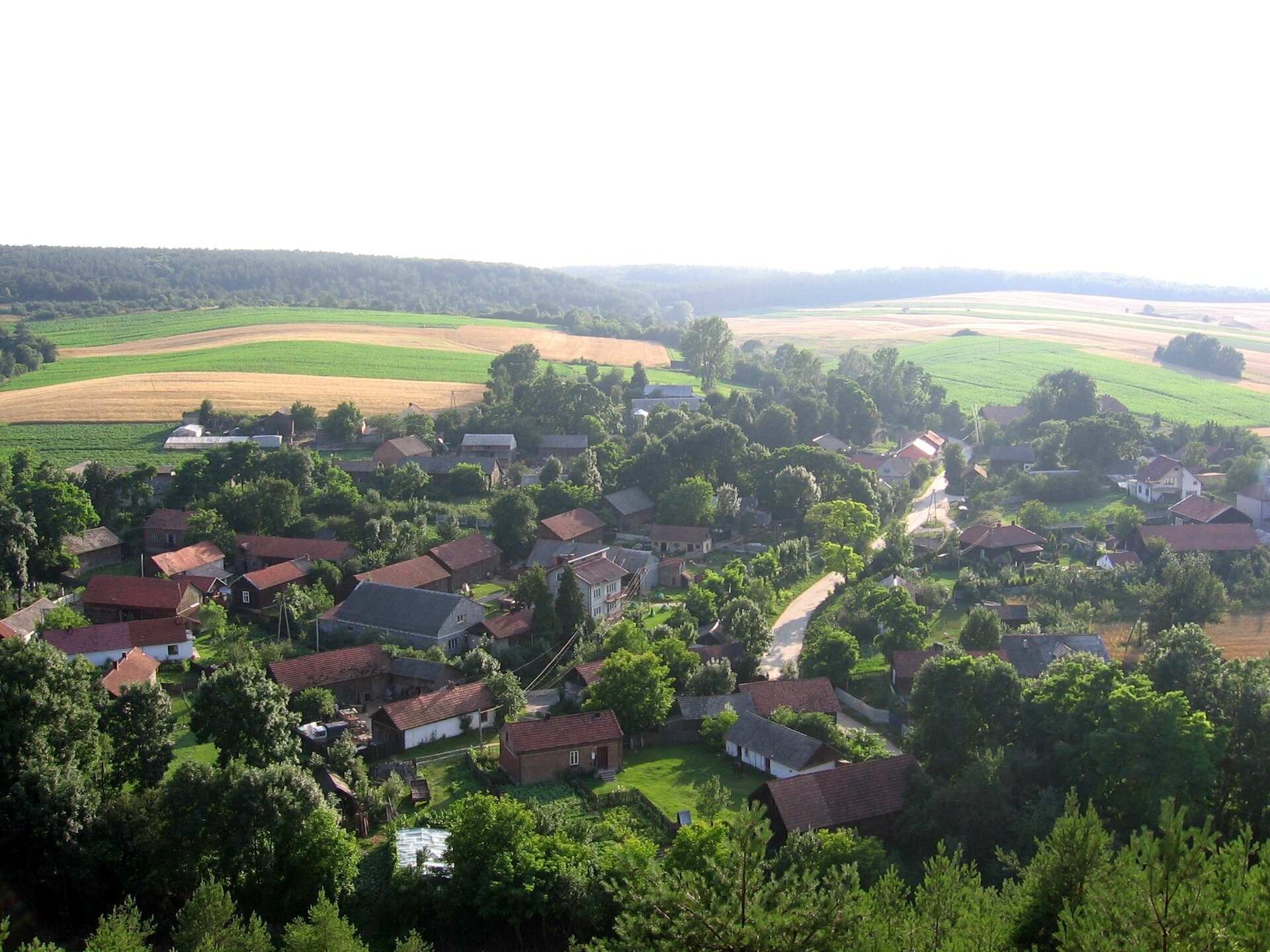 Lotnicze zdjęcie przedstawiające krętą drogę prowadzącą przez niewielką wioskę. Duża ilość małych domów jednorodzinnych. Dookoła pola, lasy. Lekko pofałdowany teren.