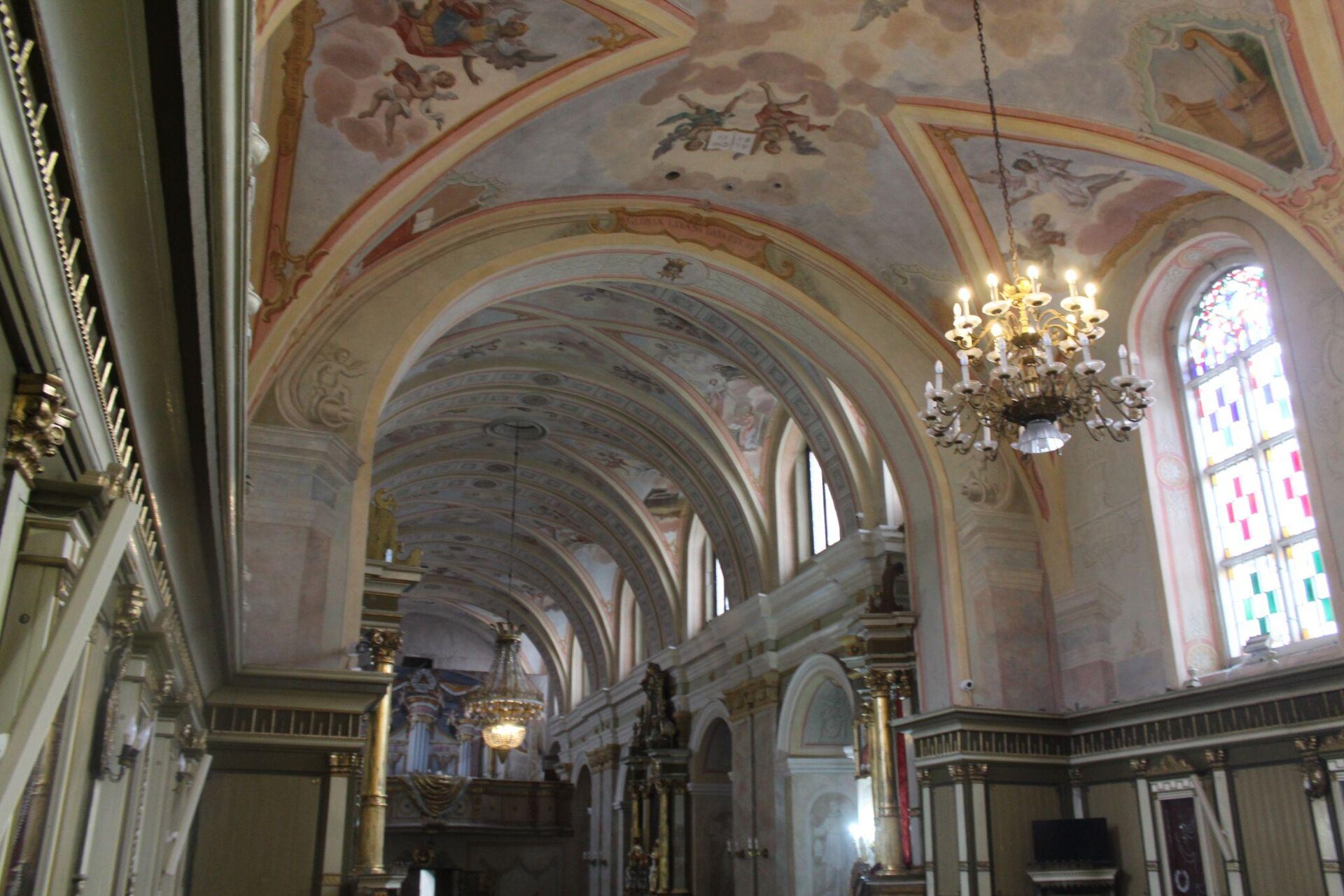 Zdjęcie przedstawia wnętrze kościoła. Sufit budynku jest bogato zdobiony malowidłami. Z sufitu zwisają dwa szklane żyrandole, które są zapalone. U dołu zdjęcia widoczne są fragmenty ścian, na których znajdują się obrazy oprawione w złote ramy.