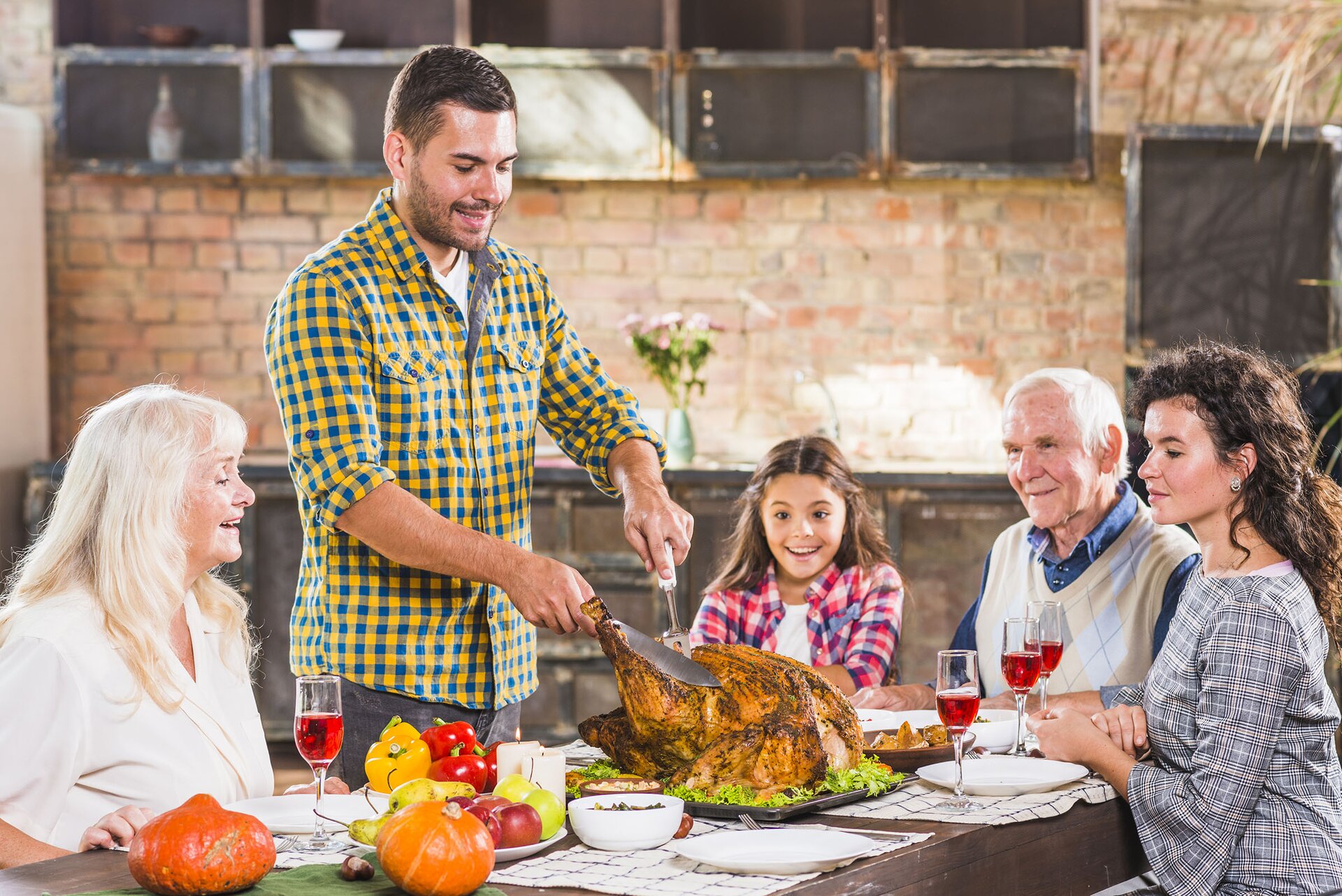 Zdjęcie przedstawia rodzinę siedzącą przy stole. widoczni są dziadkowie oraz para młodych ludzi i dziewczynka w wieku szkolnym. Młody mężczyzna kroi stojącego na środku stołu indyka. 