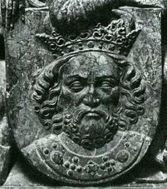 Kliknij aby powiększyć Zdjęcie przedstawia metalowy herb. W herbie widoczna jest twarz i ramiona mężczyzny w średnim wieku. Ma on kręcone włosy, a także krótką brodę. Ma zamknięte oczy. Jego nos jest duży i lekko spłaszczony na czubku. Na jego głowie znajduje się bogato zdobiona korona. Na szyi mężczyzny znajduje się gruby łańcuch, do którego przytwierdzone są ozdobne kamienie. Cały herb jest w kolorze szarym.