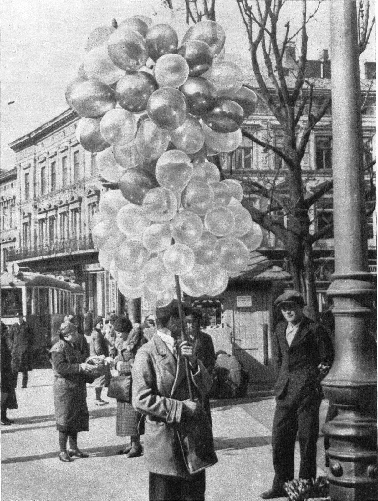 Zdjęcie przedstawia krajobraz miejski. Na pierwszym planie widać mężczyznę trzymającego mnóstwo balonów. Na drugim planie widać przekupkę, która sprzedaje coś kobiecie, przechodniów.