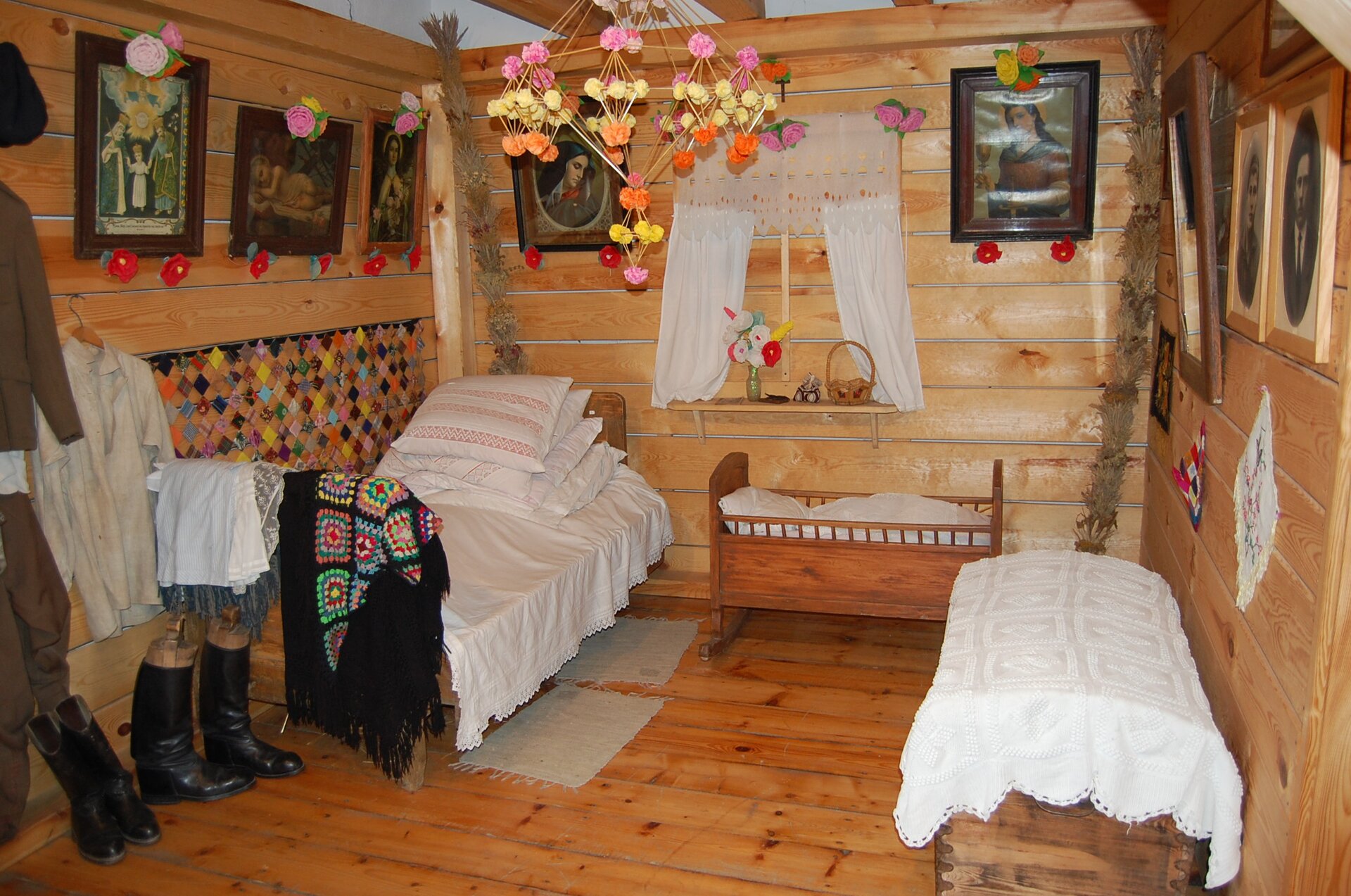Zdjęcie przedstawia wnętrze tradycyjnej wiejskiej chaty. Na zdjęciu dwa niewielkie łóżka oraz kołyska. Na ścianach wnętrza znajdują się obrazy przedstawiające religijne postacie. W lewym rogu zdjęcia widoczne są również tradycyjne stroje mieszkańców. 