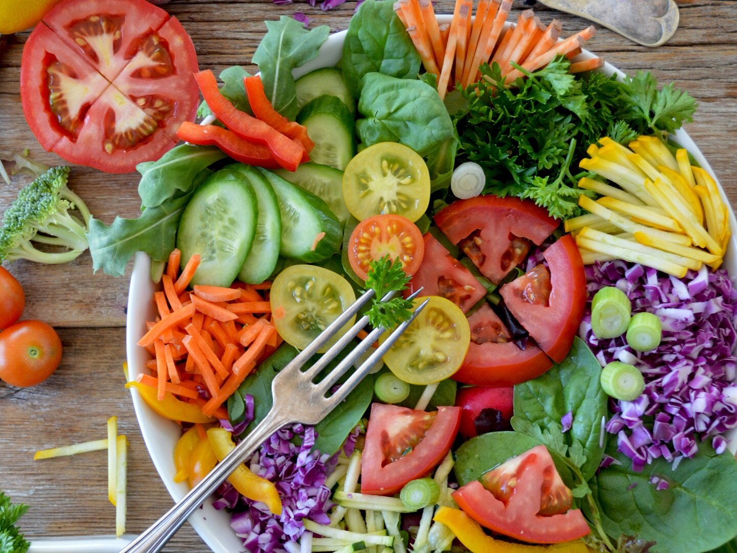 Zdjęcie przedstawia miskę pełną różnorodnych pokrojonych warzyw, takich jak pomidory, ogórki, brokuły, sałata, kapusta czerwona itp. Na misce leży widelec.