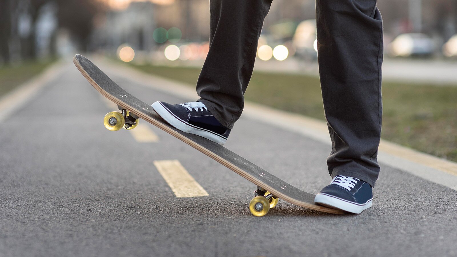 Zdjęcie przedstawia nogi stojące na deskorolce. Deskorolka jest uniesiona do góry i opiera się na dwóch kółkach. 