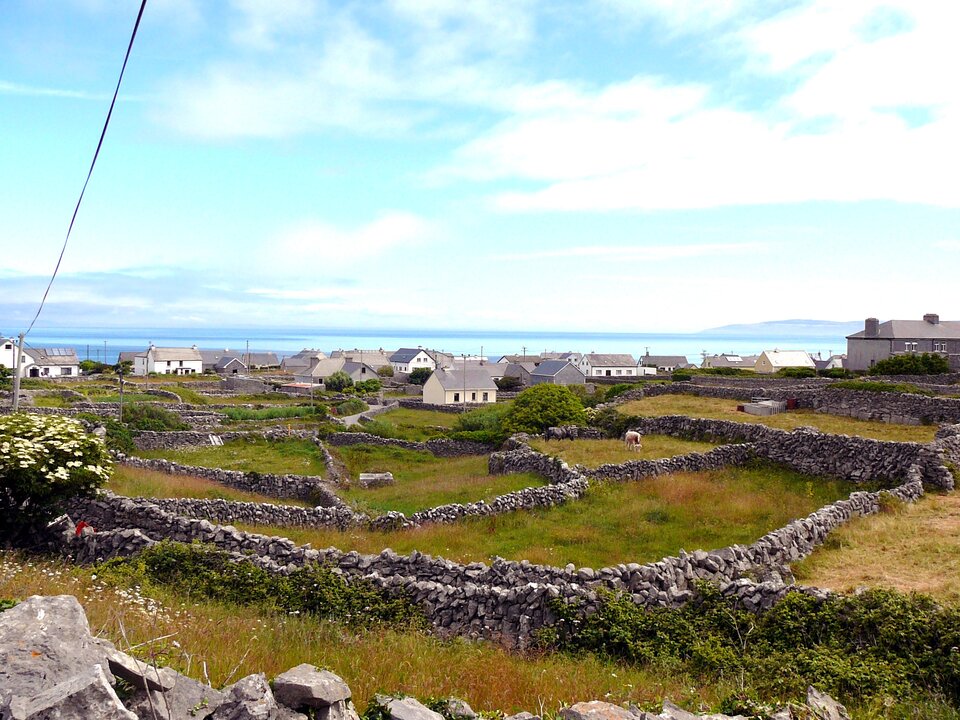 Zdjęcie przedstawia krajobraz Wysp Aran w Irlandii. Na pierwszym planie widać zagrody dla zwierząt poodzielane kamiennymi murkami. Dalej usytuowane są budynki gospodarcze i domu mieszkalne o niskiej zabudowie, w oddali morze o jasnobłękitnej barwie.