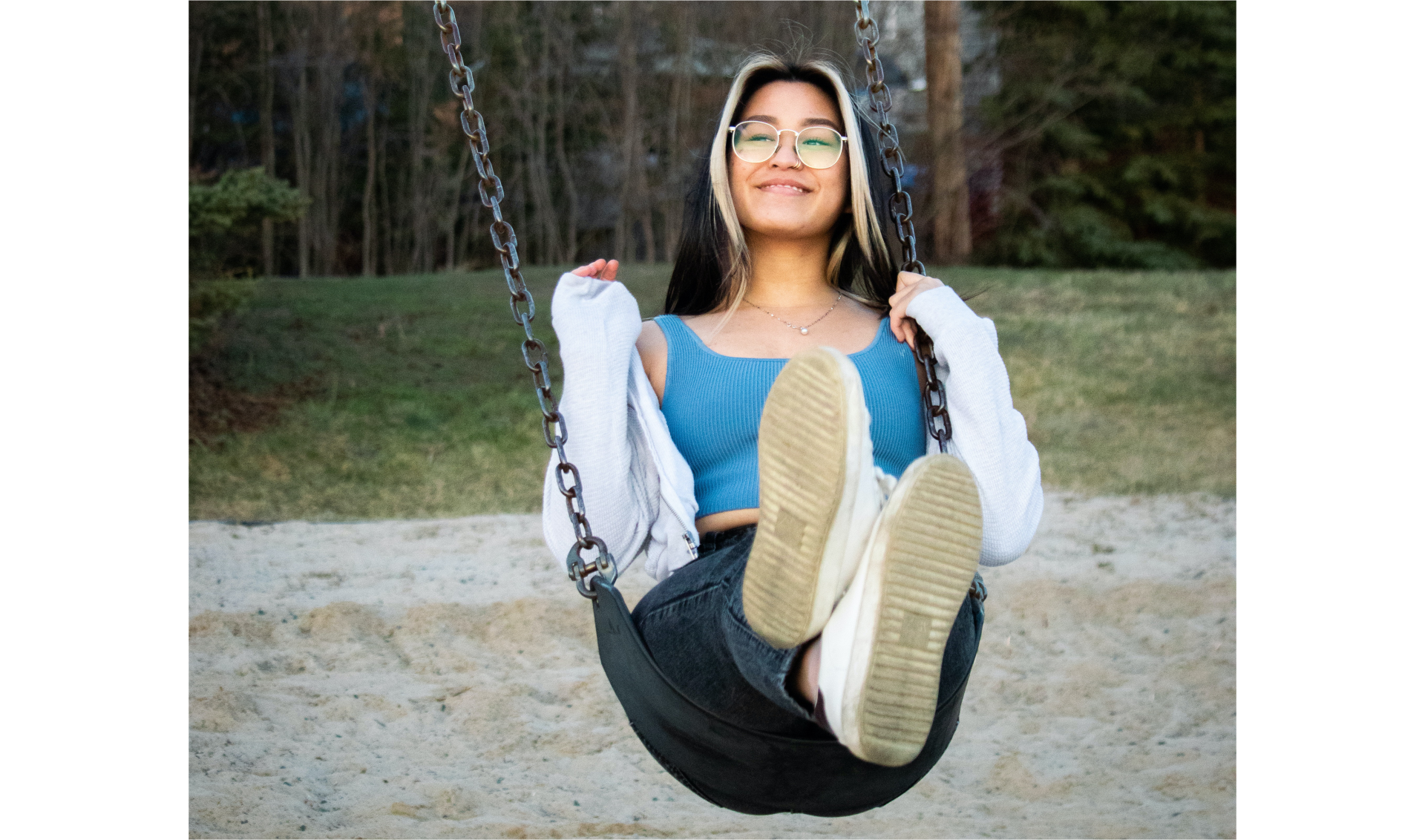 Zdjęcie dziewczyny na huśtawce w ruchu. Siedzenie huśtawki jest materiałowe, zawieszone na łańcuchach. Dziewczyna jest uśmiechnięta, ma na nosie duże okulary. W jej ciemnych włosach są blond pasemka. Na niebieski podkoszulek założoną ma białą, rozsuniętą bluzkę. Ubrana jest też w niebieskie jeansy i białe buty. W tle piasek, za nim zielone wzniesienie i dalej las.