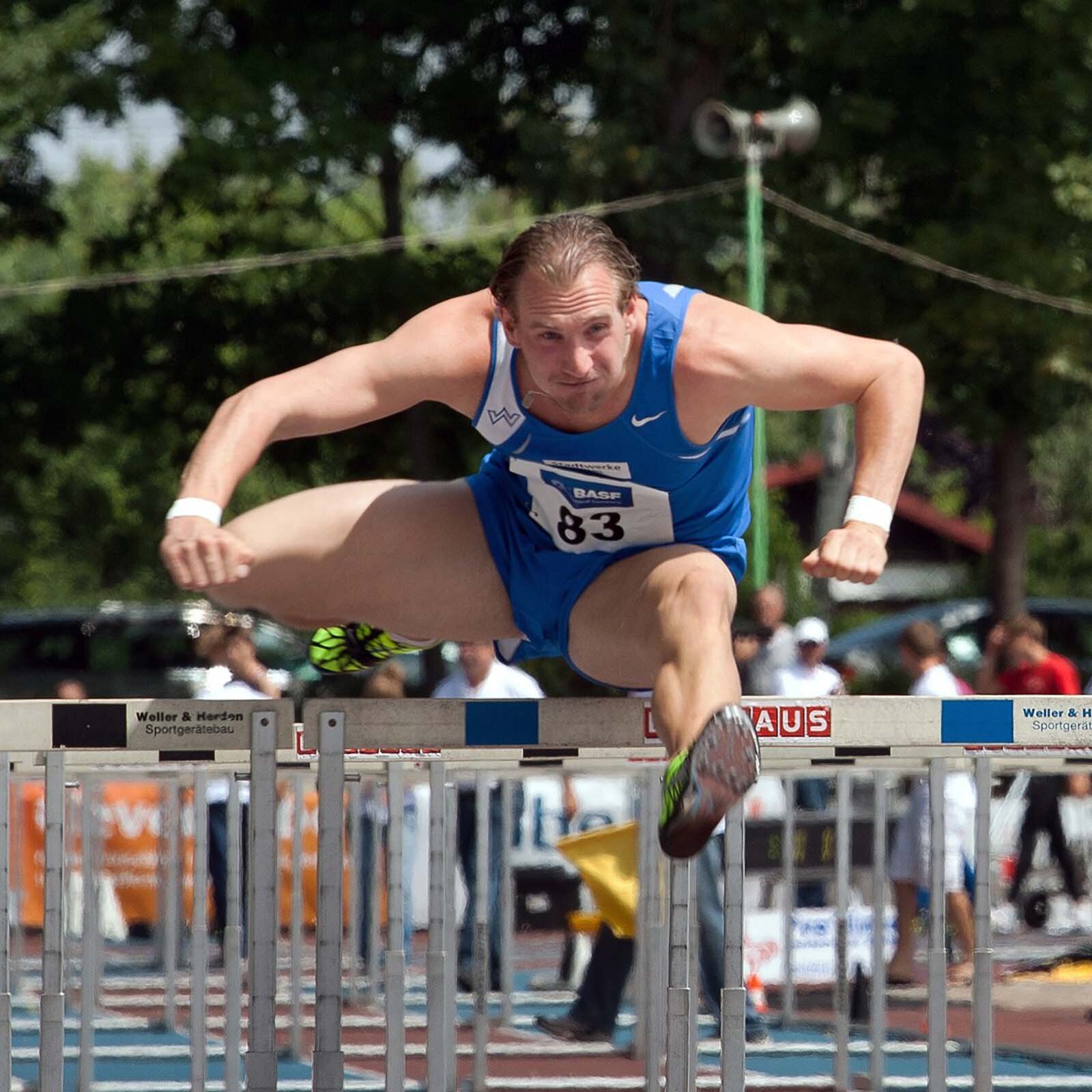 Zdjęcie przedstawia mężczyznę w stroju sportowym biegnącego przez płotki.