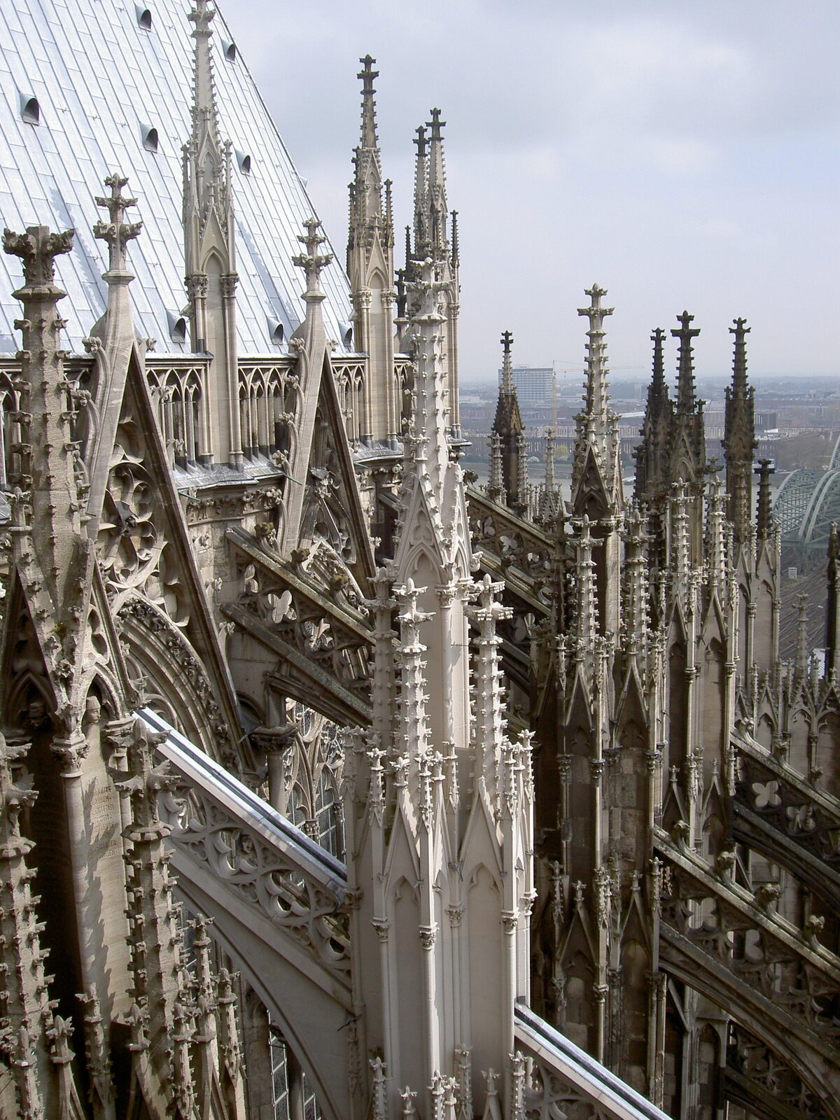 Fotografia przedstawia widok na przypory prezbiterium Katedry św. Piotra i Najświętszej Marii Panny w Kolonii w Niemczech. Na tle katedry rozciąga się zachmurzone niebo wraz z fragmentem miasta.