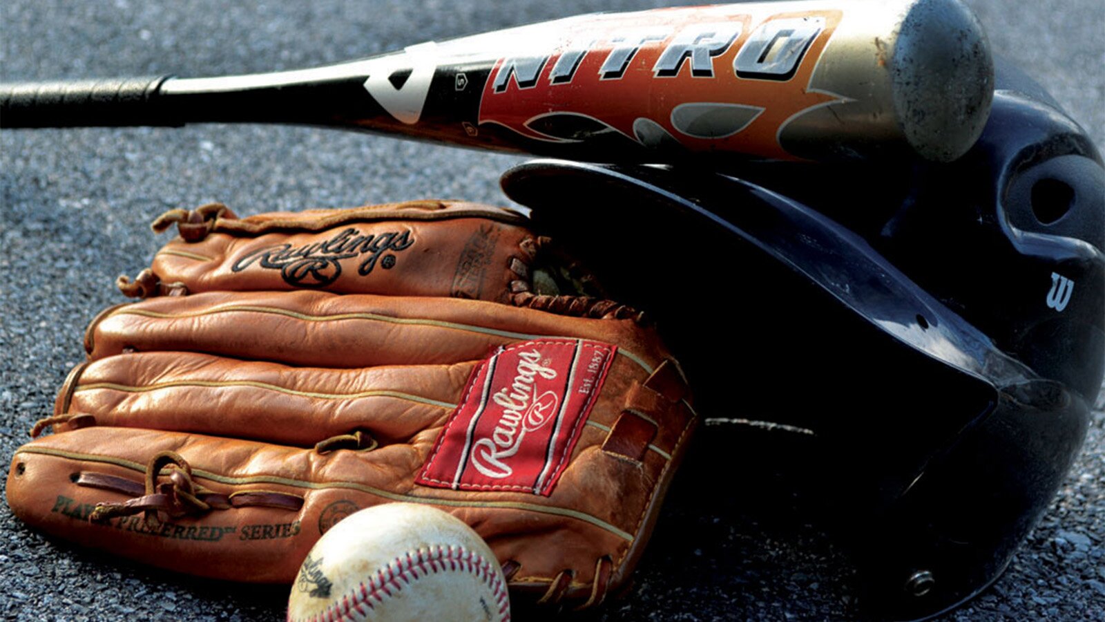 Zdjęcie przedstawia ułożone akcesoria potrzebne do gry w  baseball . Widoczne są: rękawica ochronna z brązowej skóry, kij, czarny hełm pałkarza i piłeczka. 