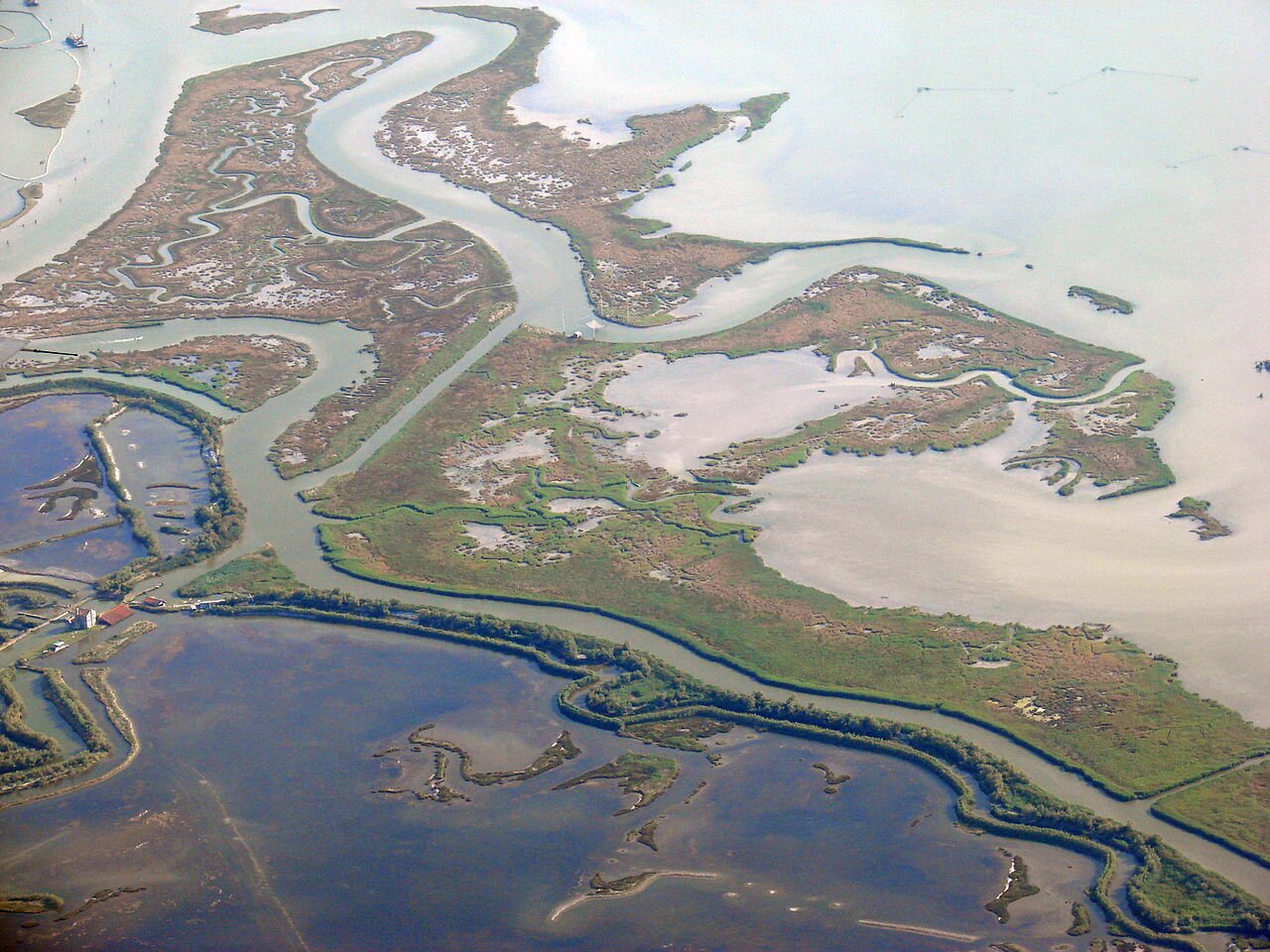 Zdjęcie przedstawia widzianą z góry Zatokę Wenecką - wybrzeże lagunowe. Widoczne zbiorniki wodne są niewielkie i przedzielone płaskimi masami ziemi, która to usunięta została w wielu miejscach, aby stworzyć kanały. 