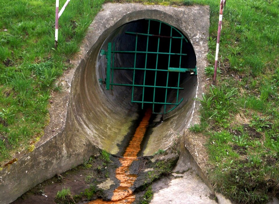 Zdjęcie przedstawia rurę wylotową kanalizacji. Z rury wypływa niewielki strumień pomarańczowych ścieków. 