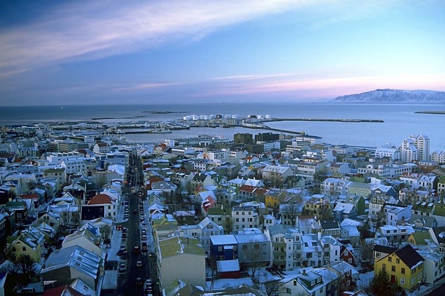 Zdjęcie przedstawia widok z góry na stolicę Islandii, Reykjavik. Przeważa tu zabudowa niska, nie ma wieżowców ani bloków. W tle pokryta śniegiem góra Esja.