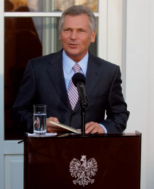Na zdjęciu znajduje się Aleksander Kwaśniewski przemawiający z małej mównicy z polskim godłem. Ma on krótkie, siwiejące włosy i jest ubrany w garnitur. W rękach trzyma małą książkę. Na mównicy znajduje się mikrofon oraz szklanka wody.