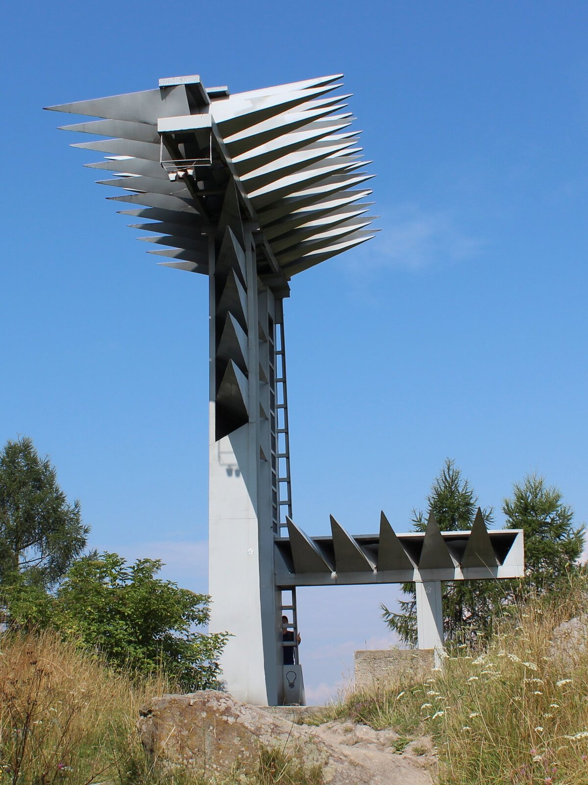 Ilustracja przedstawiająca pomnik autorstwa Władysława Hasiora pt. „Organy"(„Pomnik dla poległych w walce o utrwalenie władzy ludowej na Podhalu”). Pomnik przedstawia konstrukcję przypominającą organy, umieszczoną na słupie. Składa się z dwóch części: pionowej i poziomej. Element poziomy to betonowa płyta wystająca poza szczyt wzgórza, na którym został umieszczony pomnik. Pionowe elementy są najeżone długimi, geometrycznymi, trójkątnymi "kolcami". 