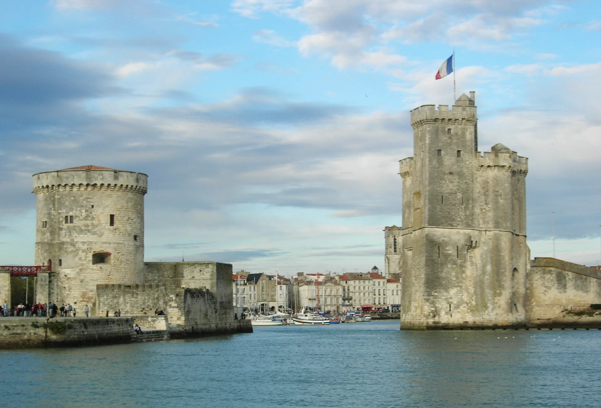 Na zdjęciu La Rochelle, wejście do portu. Widoczne dwie wieże, z lewej i prawej. Wybudowane z kamienia. Na wprost budynki miejskie. Wieże otoczone spokojną wodę.  
