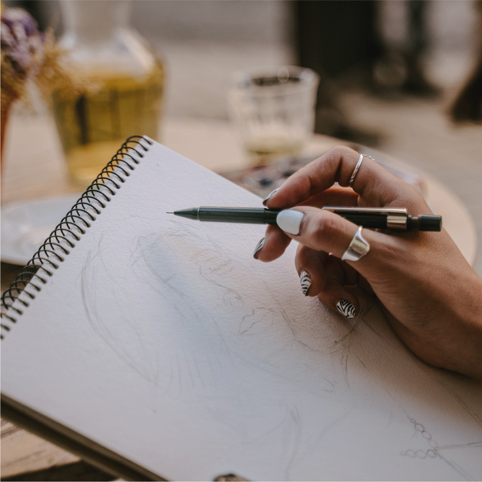 Zdjęcie przedstawia szkicownik oraz dłoń kobiety trzymającą ołówek. Na kartce szkicownika, widoczny jest szkic powstającego rysunku. Dłoń kobiety oparta jest nadgarstkiem o kartkę papieru. Kobieta nosi pierścionek na kciuku oraz palcu wskazującym. Jej paznokcie są pomalowane w różne wzory.