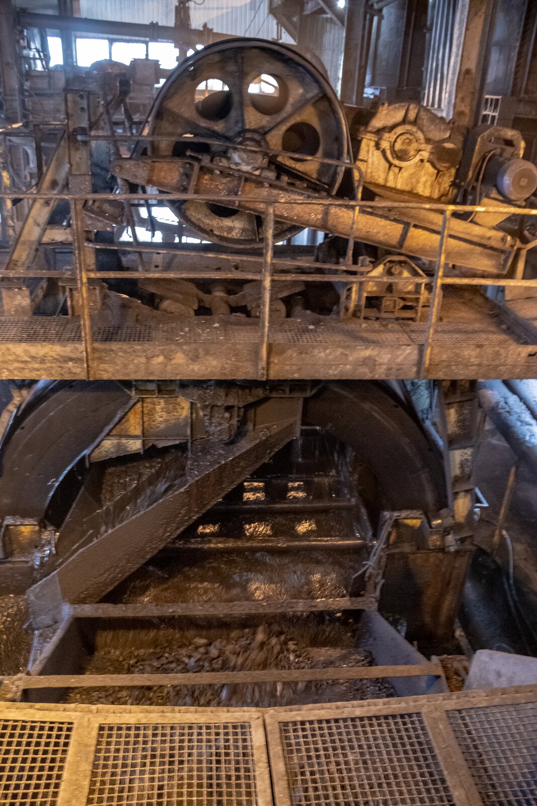 Ilustracja przedstawia model wzbogacalnika zawiesinowego typu DISA. Urządzenie ma kształt walca z usytuowanymi pionowo podstawami i niedużą wysokością ściany bocznej. Wzbogacalnik zawiesinowy typu DISA zbudowany jest ze skrzyni roboczej, czyli zbiornika, do którego doprowadzane są nadawa oraz ciecz zawiesinowa. W skrzyni roboczej dochodzi do rozdziału na dwa produkty. Zamontowane są w niej wszystkie pozostałe elementy składowe wzbogacalnika. Od spodu skrzyni przyłączone są dwa rurociągi doprowadzające ciecz ciężką. Kolejnym elementem jest zsuwnia oraz zamontowany tuż pod nią rurociąg. Umożliwiają doprowadzenie do górnej części skrzyni roboczej, odpowiednio nadawy oraz poziomym strumieniem - cieczy ciężkiej. Zsuwnia oraz rurociąg znajdują się po stronie jednej walca przy jednej z podstaw, którego kształt przypomina całe urządzenie. Przelew to część wzbogacalnika, którą odprowadzany jest koncentrat, czyli frakcja pływająca. Znajduje się po przeciwnej stronie wzbogacalnika względem zsuwni, to znaczy przy drugiej podstawie walca, którego kształtem przypomina urządzenie Tuż nad lustrem cieczy przed przelewem zamontowany jest obrotowy wygarniacz łopatkowy. Przyspiesza on wyodrębnianie koncentratu, zgarniając go z lustra cieczy za próg przelewowy koryta roboczego. To jest wyprowadza frakcję pływającą poza wzbogacalnik celem jej dalszej obróbki. Wygarniacz stanowi usytuowany poziomo, równolegle do linii przelewu trzon obracający się w trakcie pracy urządzenia wokół własnej osi. Wokół niego rozmieszczone są promieniście dookoła wspomnianej osi, na całej długości trzonu płaskie, prostokątne elementy, czyli łopatki. W środku skrzyni znajduje się koryto robocze. Jest to część wzbogacalnika, w której ma miejsce właściwy podział. To znaczy frakcja pływająca niesiona strumieniem cieczy kierowana jest do przelewu, zaś frakcja tonąca opada na dno, osiadając na kole łopatkowym. Koryto robocze oddzielone jest od skrzyni ściankami, których dolne krawędzie znajdują się tuż nad kołem łopatkowym. Wspomniane koło łopatkowe to kolejny element wzbogacalnika, pozwala na wyprowadzenie frakcji tonącej z koryta roboczego. Koło zamontowane jest wzdłuż ściany walca, do którego zbliżony jest kształtem wzbogacalnik. Jest ono zawieszone na taśmie napędowej i zamontowane we wnętrzu skrzyni roboczej oraz wyposażone w łopatki (przegrody) wyprowadzające frakcję tonącą. Łopatki te stanowią ruszty ze szczelinami, które umożliwiają swobodny przepływ cieczy podczas obrotu koła łopatkowego. Jednak one zatrzymują frakcję tonącą. Łopatki zamontowane są wzdłuż obwodu koła pod pewnym kątem tak, by możliwe było wyniesienie oddzielonego materiału do górnej części wzbogacalnika. Frakcja tonąca, zgarniana za pomocą łopatek, wynoszona jest na pewną wysokość i po przekroczeniu kąta zsypu zsuwa się do zsuwni odbiorczej. Zsuwnia odbiorcza to wydzielona część wzbogacalnika. Znajduje się w górnej części urządzenia, jednak nieco poniżej wysokości, na której zostaje przekroczony kąt zsypu. Jest ona oddzielona od koryta roboczego ściankami. Wyprowadza frakcję tonącą poza wzbogacalnik. Na samej górze wzbogacalnika, znajduje się platforma, z której możliwy jest dostęp do zamontowanego na jej wysokości silnika napędowego, na którym zaciągnięta jest taśma wprowadzająca w ruch koło łopatkowe. Poniżej grafiki znajduje się przycisk multimedialny z nagraniem audio. Treść nagrania jest zgodna z tytułem zdjęcia.