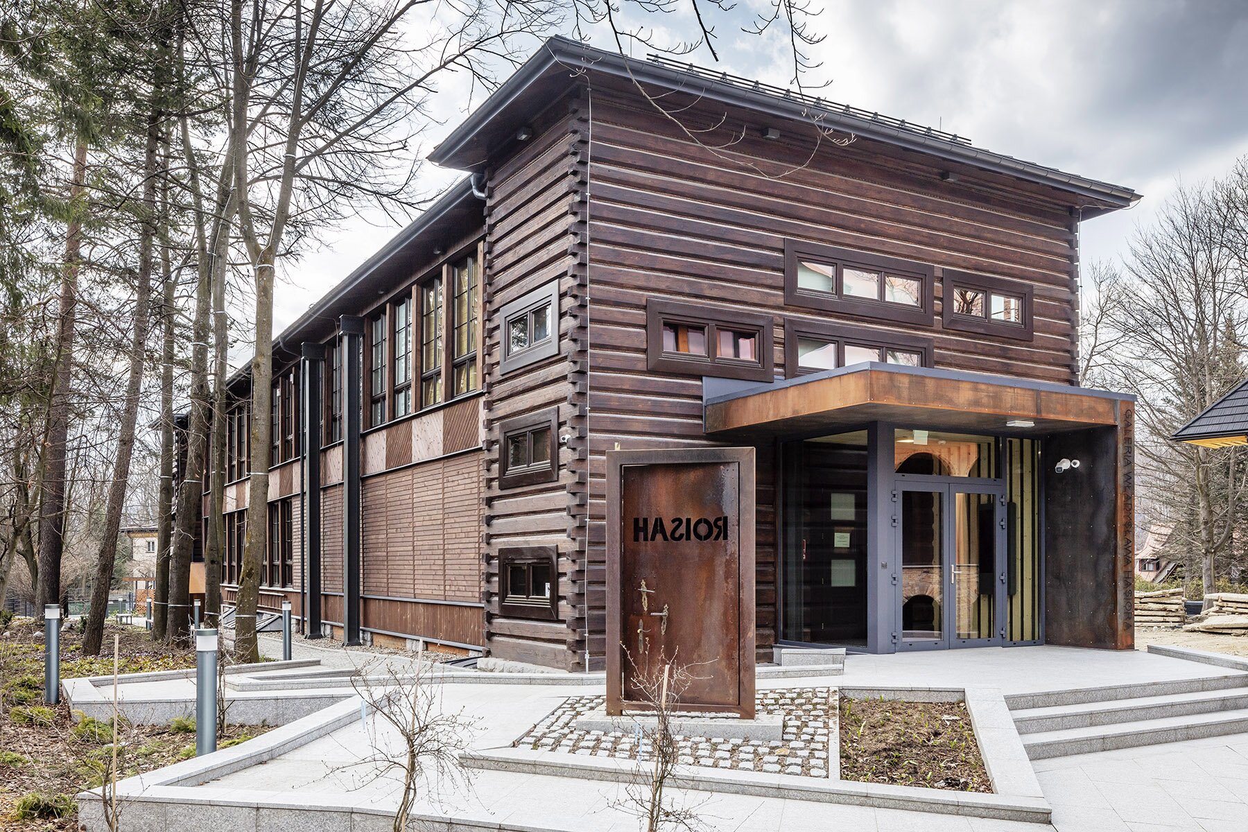 Zdjęcie przedstawia drewniany budynek w kolorze ciemnobrązowym. Z przodu znajduje się wystająca, przeszklona konstrukcja z drzwiami wejściowymi. Przed budynkiem znajduje się szary chodnik. Na nim stoją odłączone od budynku drzwi z trzema klamkami, z napisem „Hasior” z literami „s” oraz „r” przestawionymi w lustrzanym odbiciu. Dookoła rosną drzewa.