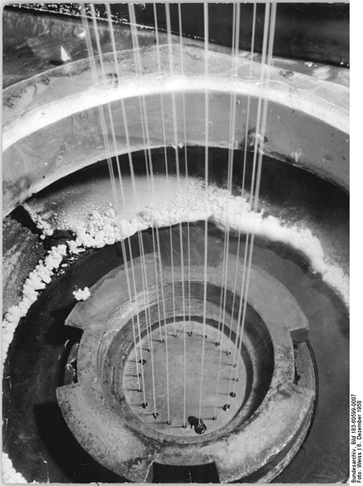 Ilustracja przedstawia proces otrzymywania wkłókien syntetycznych. Ukazuje metalowy zbiornik z małymi otworami, przez które przecieka płyn - ciekły polimer.