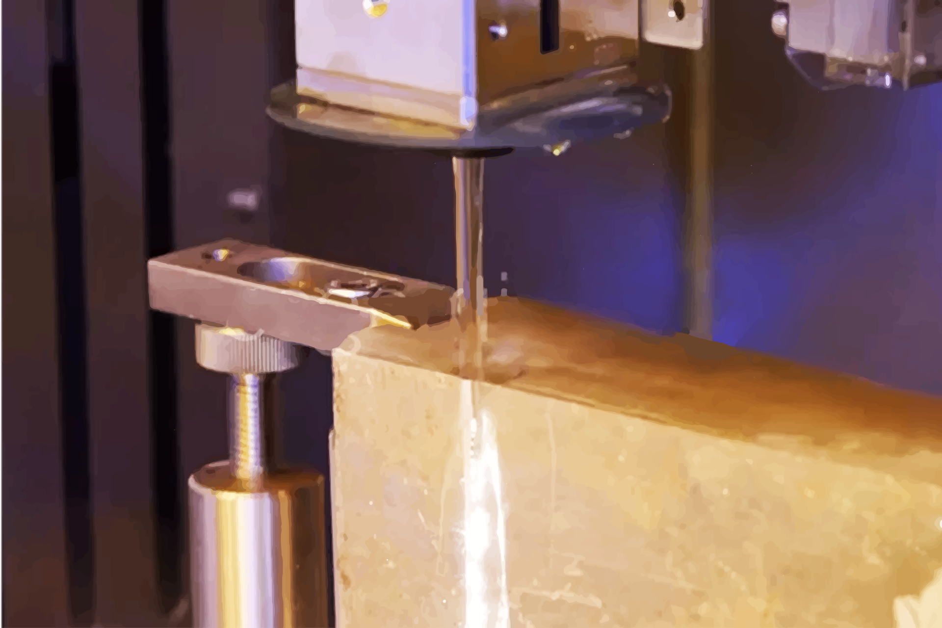 Zdjęcie ukazujące przecinanie płyty maszyną waterjet. Płyta unieruchomiona jest specjalnym zaciskiem, nad nią widoczny jest fragment urządzenia z dyszą, z której wydobywa się woda.

