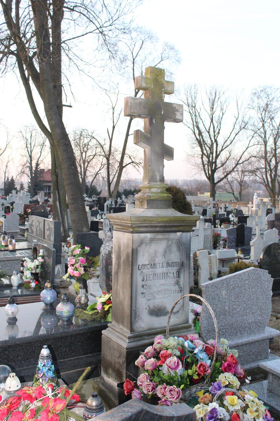 Zdjęcie przedstawia wysoki nagrobek, który zakończony jest krzyżem prawosławnym. Na nagrobku jest zamieszczona płyta pamiątkowa, na której wyryte zostały napisy cyrylicą. Na nagrobku stoją bukiety sztucznych kwiatów. W tle widoczne są kolejne, współczesne nagrobki. Pomiędzy nagrobkami rosną bezlistne drzewa. Niebo jest jasne, bezchmurne. Zdjęcie zostało wykonane w świetle dziennym.