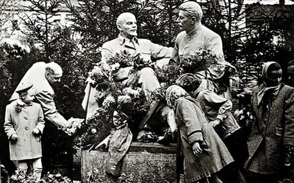 Fotografia przedstawiająca pomnik Lenina i Stalina w Krakowie, w parku Strzeleckim. Mężczyźni ukazani na poniku siedzą i rozmawiają ze sobą. Pod pomnikiem widać ludzi składających kwiaty, są wśród nich liczne dzieci.