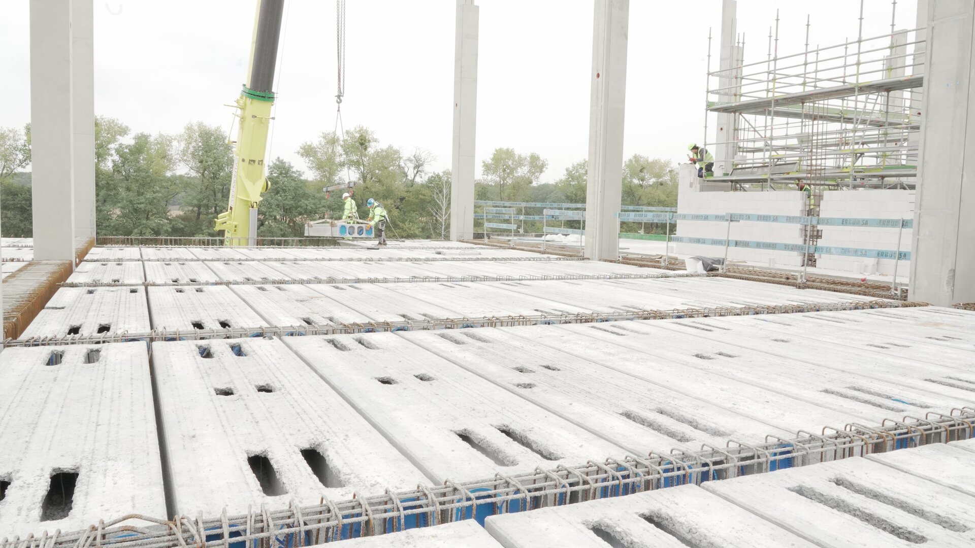 Zdjęcie przedstawia zbrojenie stropu. W głębi zdjęcia widać dwóch robotników pracujących przy ramieniu dźwigu.