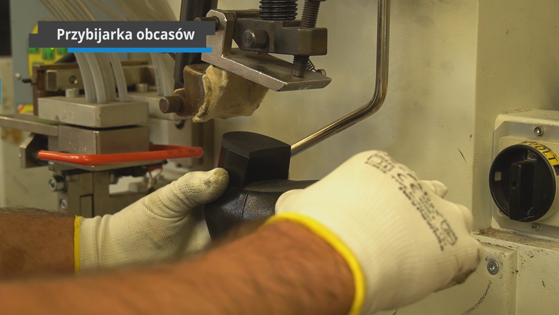 Na zdjęciu widoczne są ręce w rękawiczkach, trzymające but. Przy użyciu maszyny jest do niego przybijany obcas. Nagranie tożsame z treścią.