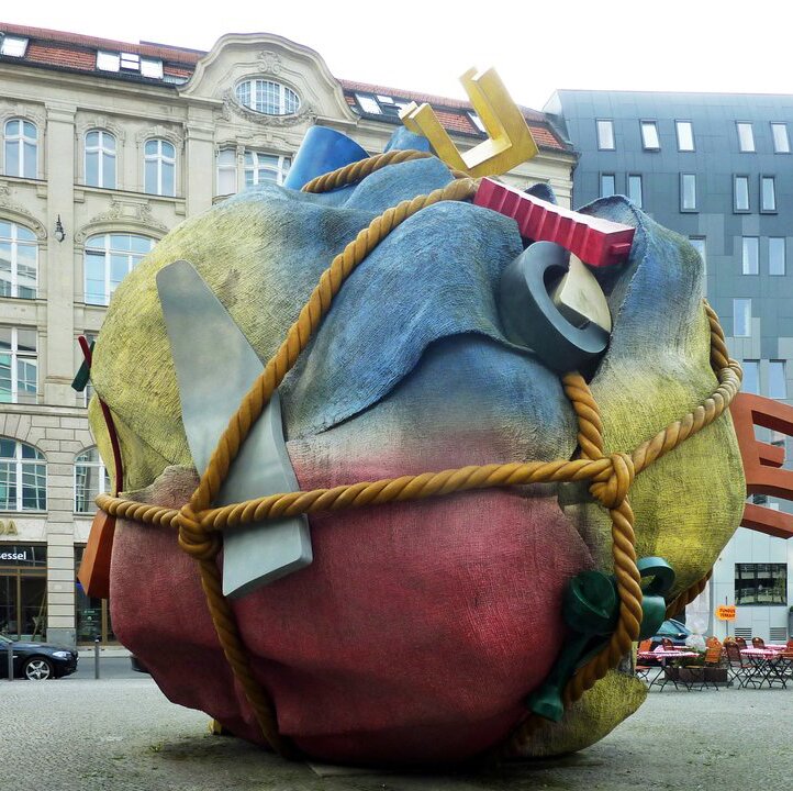 Zdjęcie przedstawia rzeźbę, która przypomina zmiętą kartkę papieru, która jest obwiązana sznurem. Rzeźba jest żółto-czerwono-niebieska. Można mieć wrażenie, że jej elementy są w nią wtopione – wśród nich opona, krzesło.