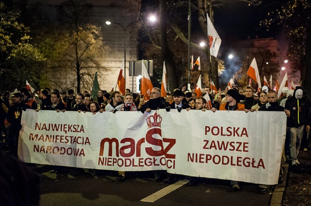 Zdjęcie przedstawia idący ulicą tłum ludzi. Widać biało-czerwone flagi i palące się race. Na czele marszu idą mężczyźni z transparentem, na którym widnieje napis: Marsz Niepodległości. Polska dla Polaków. Polacy dla Polski. Widać także transparent z napisem: Stop islamizacji.