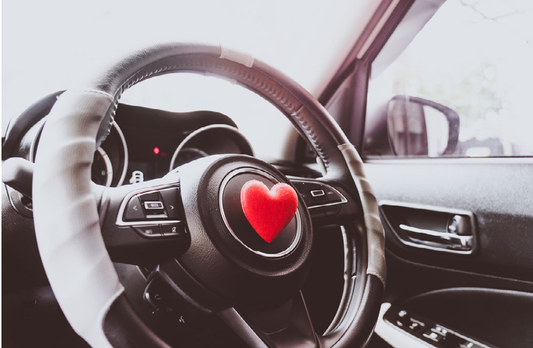 Ilustracja przedstawia fragment wnętrza samochodu w monochromatycznej kolorystyce z akcentem w postaci czerwonego serca na środku kierownicy.